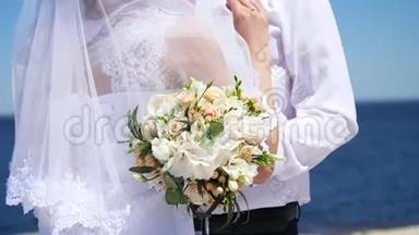 婚礼花束的特写，新婚夫妇穿着婚纱拥抱在一起。 在背景中，大海和天空